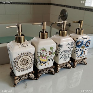 May include: Four square ceramic soap dispensers with bronze-toned pump mechanisms and ornate, decorative bronze-toned bases. Each dispenser features a unique pattern, including a central mandala, a floral motif, a bird illustration, and blue flowers.