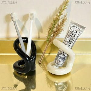 May include: A black twisted ceramic holder containing a grey and a white toothbrush, and a cream-coloured twisted ceramic holder containing a silver tube of Marvis toothpaste. The items are arranged with a sprig of dried grass between them.