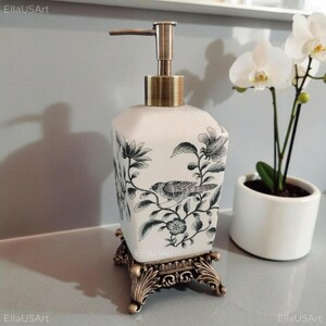 May include: A square ceramic soap dispenser featuring a black and white botanical print with a bird perched on a branch. The dispenser has a metallic bronze-toned pump mechanism and rests on an ornate, four-legged bronze-toned metal base.