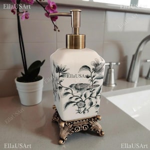 May include: A square white ceramic soap dispenser featuring a black botanical print of a bird perched on a branch. The dispenser has a metallic bronze-toned pump mechanism and rests on an ornate, four-legged bronze-toned metal base stand.