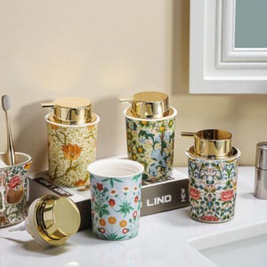 Könnte beinhalten: Eine Sammlung von Badezimmeraccessoires mit floralen Mustern, darunter Seifenspender mit goldfarbenen Deckeln, ein Zahnbürstenhalter und ein Becher. Die Designs zeigen verschiedene Blumenmuster in Grün-, Blau- und Orangetönen. Die Accessoires sind auf einer weißen Oberfläche angeordnet.