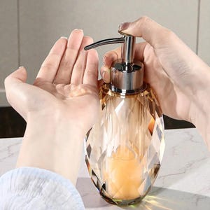 May include: A decorative, faceted glass soap dispenser with a silver pump. The dispenser is filled with a light orange liquid. The dispenser is being used to dispense soap into a person's hand.