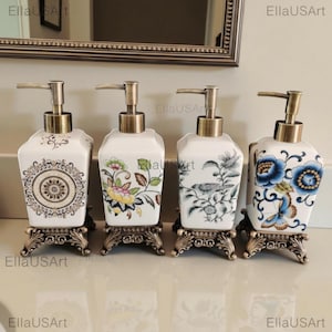 May include: Four square ceramic soap dispensers with antique bronze-toned pump mechanisms and ornate, decorative metal bases. Each dispenser features a unique pattern: a central mandala, colorful floral vines, a bird on a branch, and blue flowers with a butterfly.