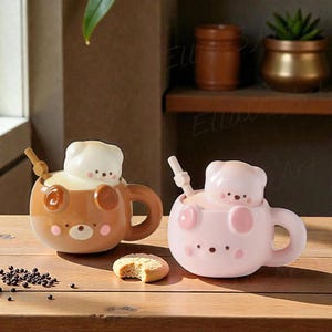 May include: Two ceramic mugs shaped like bears, one brown and one light pink, each featuring a sculpted bear lid, a matching handle, and a small stirring spoon. A single round cookie sits on the surface between the two mugs, with small dark sprinkles scattered nearby.