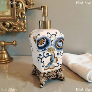 May include: A white ceramic soap dispenser with a square base, featuring blue floral patterns and a butterfly design. It has a metallic bronze-toned pump mechanism and sits on an ornate, decorative bronze-colored metal stand with four feet.