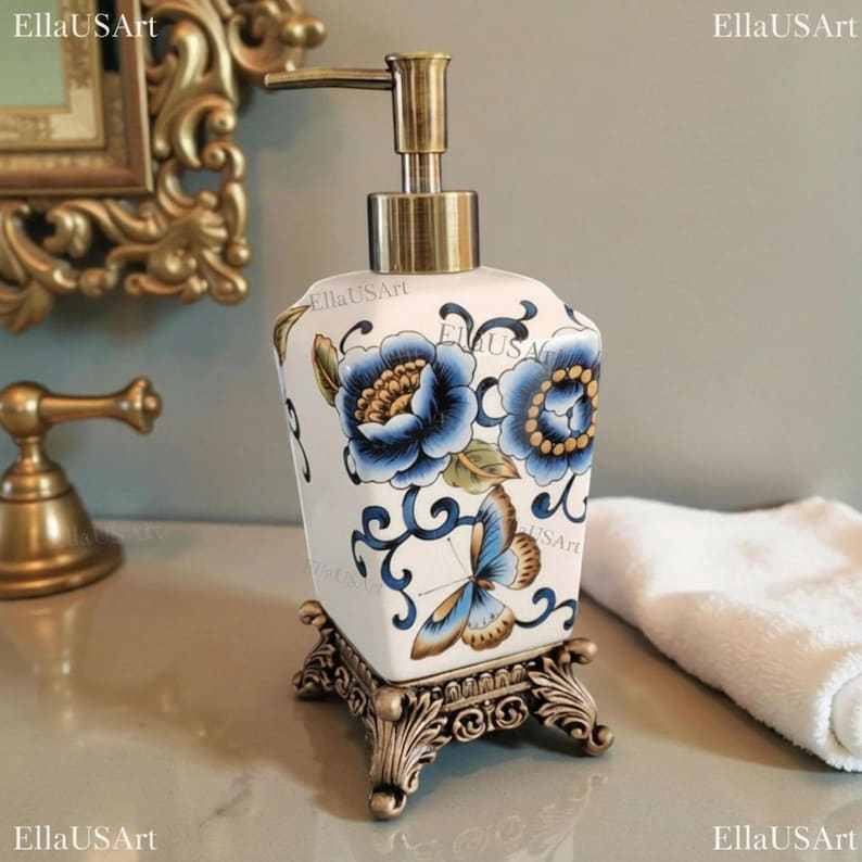 May include: A white ceramic soap dispenser with a square base, featuring blue floral patterns and a butterfly design. It has a metallic bronze-toned pump mechanism and sits on an ornate, decorative bronze-colored metal stand with four feet.