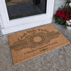May include: A rectangular doormat made of natural fibers. The mat features a Celtic knot design in the center, with the words "Céad Míle Fáilte" and "A Hundred Thousand Welcomes" in black lettering. Green accents are also present.
