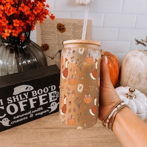 May include: A frosted glass tumbler with a bamboo lid and clear straw, featuring a repeating pattern of geese wearing orange sweaters, pumpkins, small bouquets, and books on a tan background. The glass has a cylindrical shape and smooth finish.