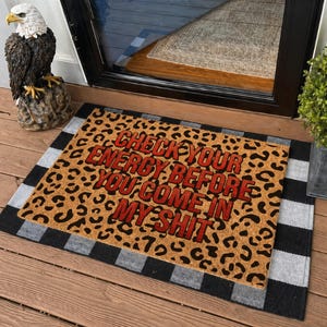 May include: A rectangular doormat with a leopard print background and the text "CHECK YOUR ENERGY BEFORE YOU COME IN MY SHIT" in red. The mat has a black and white checkered border.