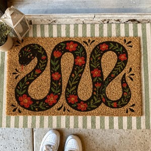 Peut inclure: Un paillasson rectangulaire avec un motif de serpent noir orné de fleurs rouges et de feuilles vertes. Le tapis est en fibre naturelle et repose sur un tapis à rayures vertes et blanches. Des baskets blanches sont visibles au premier plan.