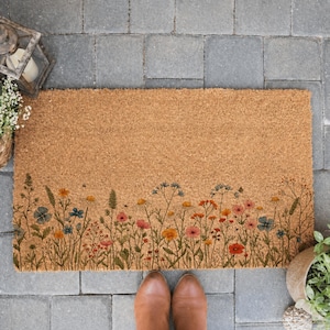 May include: A rectangular doormat made of natural coir fiber. The mat features a colorful floral border with wildflowers in shades of pink, orange, blue, and yellow. Brown boots are visible at the bottom of the mat.