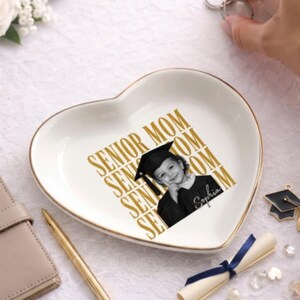 Puede incluir: Un plato decorativo de cerámica blanca en forma de corazón con borde dorado. El centro presenta el texto dorado repetido "SENIOR MOM" y una fotografía en blanco y negro de una persona graduada con birrete y toga con el nombre "Sophia".