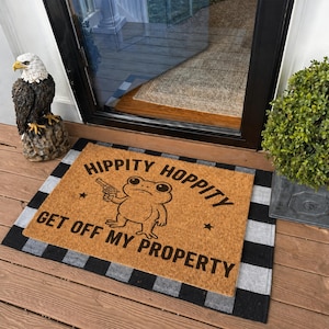 Może przedstawiać: Wycieraczka z włókna kokosowego z żabą trzymającą pistolet i napisem "HIPPITY HOPPITY GET OFF MY PROPERTY". Mata ma brązowe tło i czarno-szarą obramówkę w kratę.