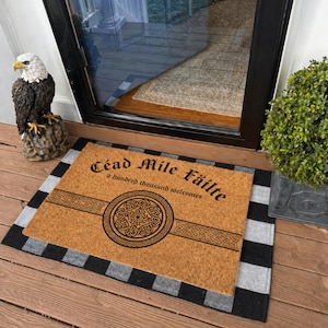 Può includere: Un zerbino rettangolare con il testo "Céad Míle Fáilte" e "a hundred thousand welcomes" in nero. Il tappeto ha un centro marrone con un disegno a nodo celtico e un bordo a strisce nere e grigie. Una statua di aquila è a sinistra.