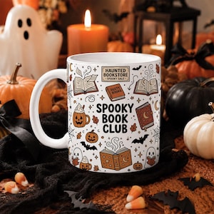 May include: A white ceramic mug featuring a black and orange Halloween-themed illustration. The design includes the text "HAUNTED BOOKSTORE SPOOKY SALE" and "SPOOKY BOOK CLUB" surrounded by drawings of open books, pumpkins, ghosts, bats, and spiderwebs.