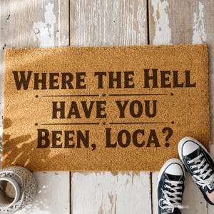 Peut inclure: Un paillasson rectangulaire avec le texte "WHERE THE HELL HAVE YOU BEEN, LOCA?" en marron foncé. Le tapis est de couleur marron clair et est placé sur une surface en bois blanc. Des baskets noires sont visibles.