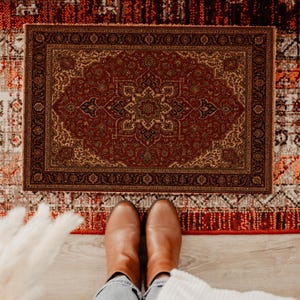 May include: A rectangular, ornate rug with a central medallion design in shades of red, gold, and black. The rug features a detailed border and is placed on a wooden floor. The image also shows a pair of brown leather boots.