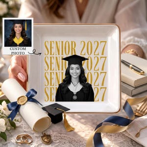 Puede incluir: Un plato cuadrado de cerámica blanca con borde dorado que presenta el texto SENIOR 2027 en letras doradas y un retrato en blanco y negro de un graduado con birrete y toga. El plato se muestra junto a un diploma enrollado con una cinta azul.