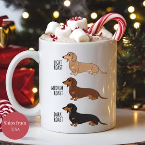 May include: White ceramic mug with illustrations of dachshunds in light, medium, and dark brown shades. The mug contains hot chocolate, marshmallows, and a candy cane. Text on the mug reads "Light Roast", "Medium Roast", and "Dark Roast".