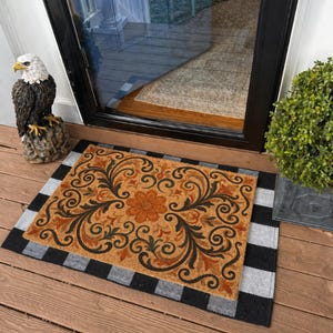 May include: A rectangular doormat featuring a floral and scroll pattern in brown, black, and orange. It has a black and grey chequered border. The mat is positioned in front of a glass door.