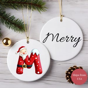 Puede incluir: Adorno navideño de cerámica blanca con una cuerda dorada. Un adorno presenta la letra "M" roja decorada como Papá Noel. El otro adorno tiene la palabra "Merry" en escritura negra. Una decoración festiva.