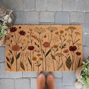 May include: A rectangular doormat with a floral design. The mat features a natural coir fiber background with a botanical illustration of various flowers in shades of burgundy, peach, and gold. The design includes roses, poppies, and other wildflowers.
