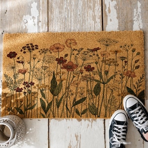 May include: A rectangular doormat with a floral design. The mat features a tan background with a detailed illustration of wildflowers in shades of red, orange, and green. Black canvas sneakers with white soles and laces are visible in the bottom right corner.