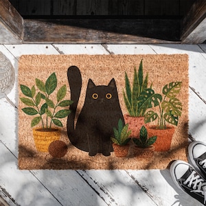 Könnte beinhalten: Rechteckige Fußmatte aus Naturfasern mit einer Illustration einer schwarzen Katze, umgeben von Topfpflanzen. Die Katze hat gelbe Augen und sitzt aufrecht. Ein Wollknäuel liegt in der Nähe der Katze. Schwarz-weiße Turnschuhe sind zu sehen.