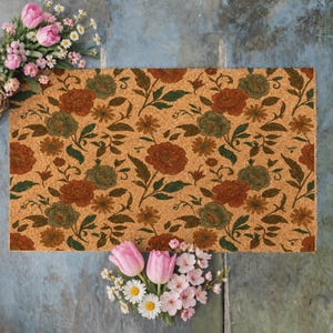 Puede incluir: Un felpudo rectangular de fibra de coco con un patrón floral repetitivo de grandes flores de color óxido y verde apagado, hojas verde oscuro y pequeñas flores amarillas. El felpudo está rodeado de tulipanes rosas y margaritas blancas.