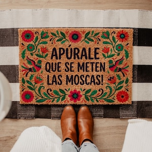 May include: A rectangular doormat with the text "APÚRALE QUE SE METEN LAS MOSCAS!" in black. The mat is brown with a floral and bird design in red, green, and pink. The mat is on a black and white checkered rug.