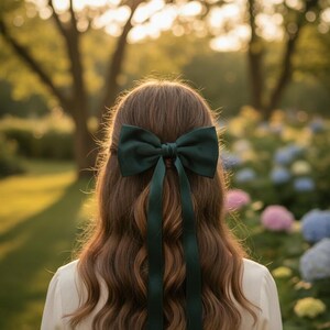 May include: A dark green bow hair accessory with long, flowing ribbons is the focal point of this image. The bow is set against the backdrop of long, wavy brown hair. The background features a blurred garden scene with trees and colorful flowers.