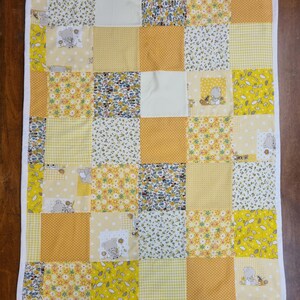 Può includere: Una trapunta patchwork per bambini con bordo bianco. La trapunta presenta quadrati di tessuti gialli, bianchi e floreali. Alcuni quadrati hanno un disegno di orsacchiotto. La trapunta è realizzata in tessuto morbido ed è adatta a un bambino.