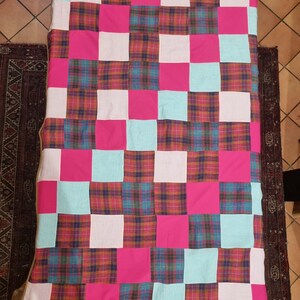 Può includere: Una trapunta patchwork con un motivo a griglia di quadrati rosa, azzurri, bianchi e a quadri. La trapunta è appoggiata su una superficie di legno, con un tappeto scuro su entrambi i lati.