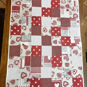 Può includere: Una trapunta patchwork con un bordo bianco, con un motivo di quadrati rossi e bianchi. I quadrati mostrano cuori, stelle, pois e altri disegni. La trapunta è realizzata con vari tessuti e trame, creando un oggetto accogliente e decorativo.