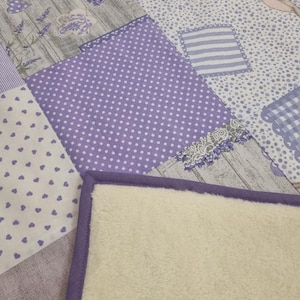 Può includere: Una trapunta patchwork con un lato inferiore morbido color crema e un bordo lavanda. La parte superiore presenta vari quadrati a motivi nei toni del lavanda, bianco e grigio, tra cui pois, cuori e disegni floreali. La trapunta ha un'estetica accogliente e fatta a mano.