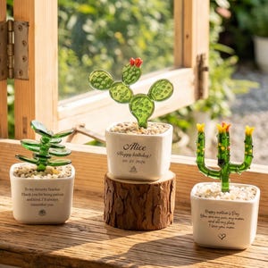 Puede incluir: Tres figuras de cactus de cristal en macetas de cerámica cuadradas blancas con piedras pequeñas. La maceta izquierda tiene un mensaje de agradecimiento. La central, sobre un tronco, dice Alice Happy birthday 09.21.2026. La derecha dice Happy mother's day.