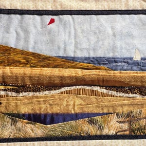 May include: A textile artwork showing a beach scene with a red kite and a sailboat. The piece uses layered fabrics in brown, tan, blue, and green. The artwork evokes a coastal landscape with a textured, layered appearance.