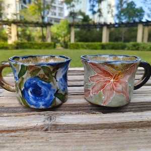 May include: Two handmade ceramic mugs with floral designs. One has blue roses, the other a red and pink flower. Both have brown handles and rims, set on a weathered wooden surface.