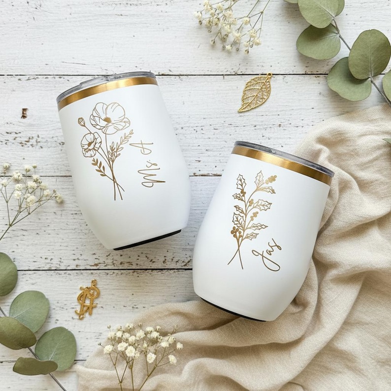 May include: Two white insulated wine tumblers with gold rims and floral designs. One tumbler has "Ms A" and the other has "Jay" in gold script. The tumblers are on a white wooden surface.
