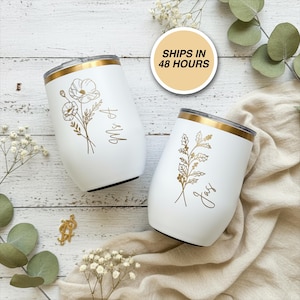 May include: Two white insulated wine tumblers with gold rims and floral designs. One tumbler has "Mrs A" and the other has "Jay" in gold script. The tumblers are on a wooden surface with floral accents.