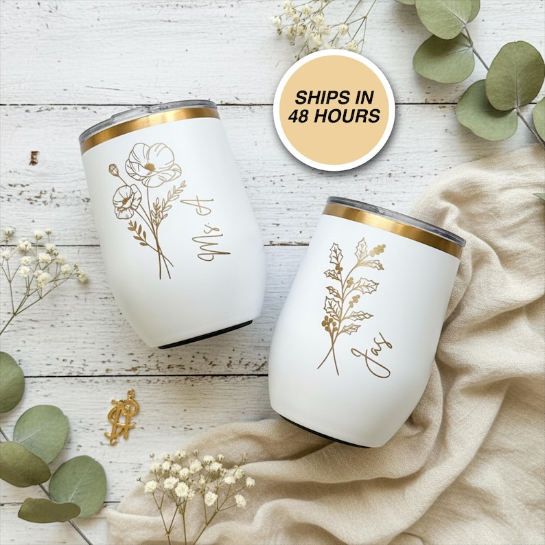 May include: Two white insulated wine tumblers with gold rims and floral designs. One tumbler has "Mrs A" and the other has "Jay" in gold script. The tumblers are on a wooden surface with floral accents.