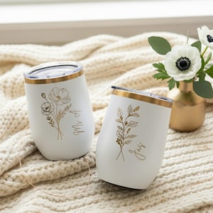 May include: Two white insulated wine tumblers with gold rims and floral designs. One tumbler has a poppy design and the text "Ms A", the other has a holly design and the text "Jas". The tumblers are on a cream-colored blanket.