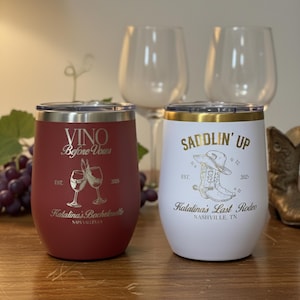 May include: Two insulated wine tumblers. One is red with the text "VINO Before Vous" and a wine glass graphic. The other is white with "SADDLIN' UP" and a cowboy boot graphic. Both have metallic silver lids.