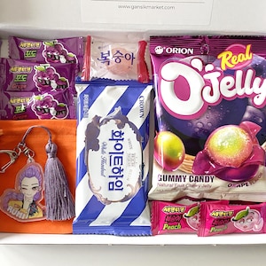 May include: A purple gift box filled with Korean snacks. The box contains grape and peach-flavored candies, a white chocolate bar, and a bag of grape-flavored jelly candies. An orange pouch with a keychain and tassel is also included.
