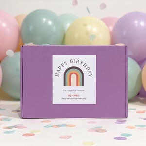 May include: A lilac gift box with a label saying "HAPPY BIRTHDAY" and "To a Special Person". The box is set against a backdrop of pastel balloons and scattered confetti.