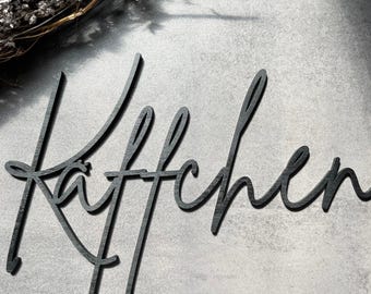 May include: A black wooden sign with the word "Kitchen?" in a cursive font. The sign is placed on a gray surface, with a sprig of dried flowers in the upper left corner.