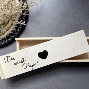 Könnte beinhalten: Eine hellfarbene Holzbox mit Schiebedeckel. Der Deckel trägt den Text "Du wirst Papa!" und ein schwarzes Herz. Die Box ist geöffnet und gibt den Blick ins Innere frei.