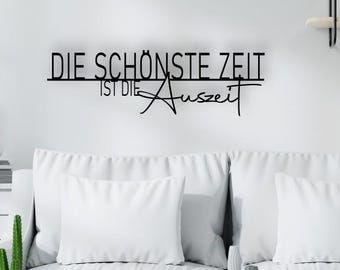 May include: A black metal wall sign featuring the German text "DIE SCHÖNSTE ZEIT IST DIE Auszeit" mounted on a white wall above a white sofa. The sign consists of thin, dark lettering in a combination of block and cursive script styles.