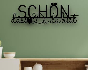 May include: A black metal wall art piece with the German phrase "SCHÖN dass Du da bist" (Nice that you are here) is mounted on a green wall. Below, a white and wood-toned cabinet holds decor.