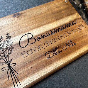 Könnte beinhalten: Ein Holzschneidebrett mit floralem Design und deutschem Text. Der Text lautet "Bonusmama, schön, dass es Dich gibt, Dein Nils." Das Brett hat eine natürliche Holzmaserung und eine dunkle Blumenillustration auf der linken Seite.